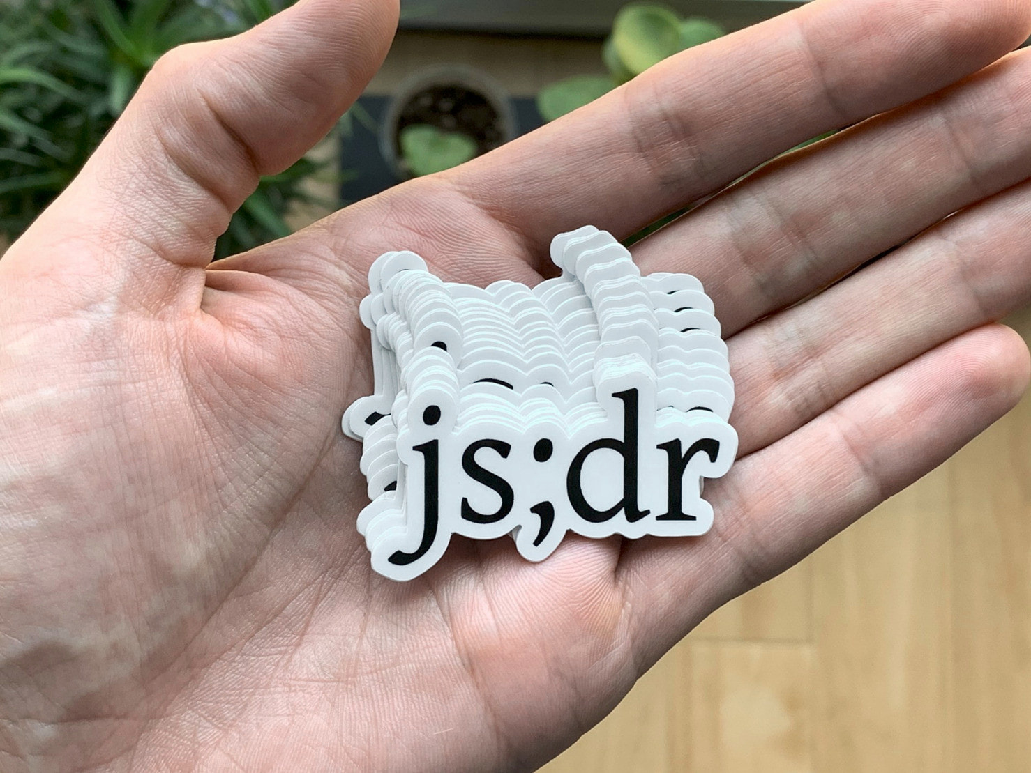 A hand holding about a dozen stickers with the “js;dr” in black on white text die-cut around the edges of the lettering