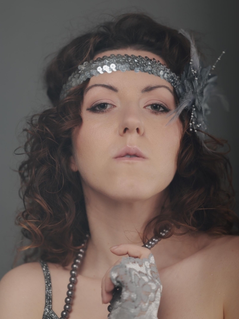 The image shows a person wearing a vintage or retro style headband with sequins and a feather. Their outfit includes a lace glove and a beaded necklace, possibly giving off a 1920s or flapper-style vibe. The person has curly hair and is looking directly at the camera.