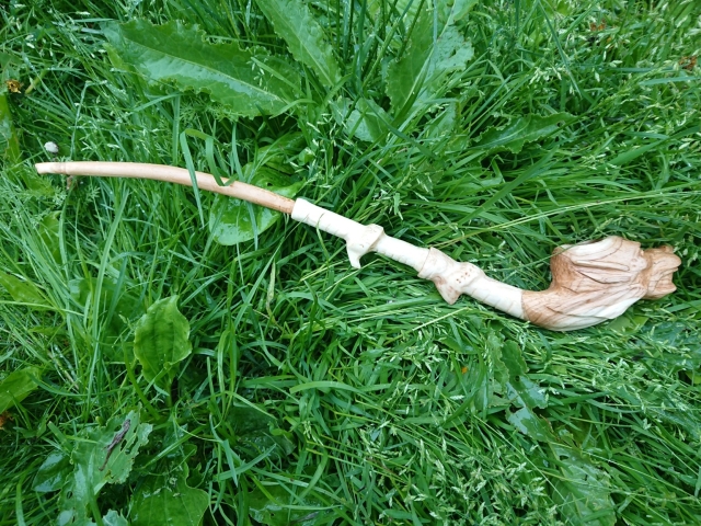 Green foliage at back round, then there's long stem wood pipe on grass . Bowl looks like dragon head. Hand carved from wood