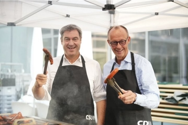 CSU Soeder und CDU Merz halten uns grinsend zwei braune Würste entgegen.