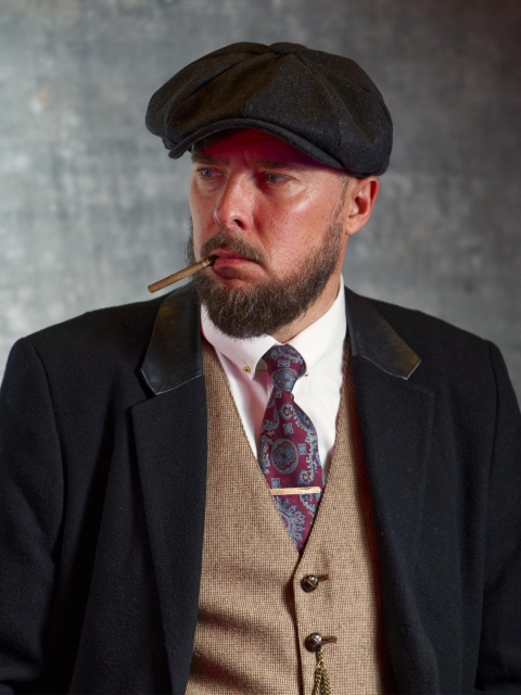 The image shows a man wearing vintage-style clothing, including a flat cap, suit jacket, vest, dress shirt, and tie. He has a beard and is holding a cigar in his mouth. The style of his clothing suggests a period look, reminiscent of early 20th-century fashion, Peaky Blinders inspired.