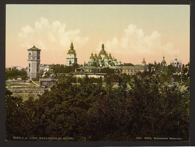 This image features an old photograph of a historic site known as Kiev Monastery St. Michael, located in the city that was formerly called Kyiv and is now part of Ukraine. The monastery complex includes several distinctive domed buildings with intricate architectural details indicative of Eastern Orthodox Christian religious architecture from its era.

The sky above has soft pinkish hues suggesting either dawn or dusk lighting conditions. In the foreground, there are dense green foliage areas which provide a natural contrast to the built environment in the background. A prominent feature is a tall tower-like structure on the left side with a conical roof and what appears to be an observation deck.

The overall image has aged characteristics such as discoloration and fading of colors that suggest it's from approximately 1900, capturing the ambiance and grandeur of historical architecture in Eastern Europe during this period.