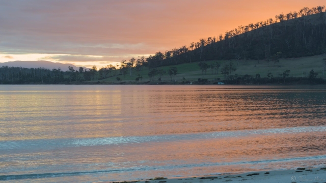 View from a beach at sunset, steep sided hill at right with trees silhouetted on the horizon, distant mountain at left.  The sky is mostly cloudy, a golden glow appearing behind the hill at right, reflected in the calm, glassy waters.