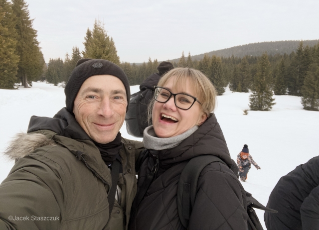 Uśmiechnięta para pozuje do selfie w śnieżnym krajobrazie, otoczona drzewami iglastymi. Kobieta, ubrana w okulary i czarną kurtkę, śmieje się, podczas gdy mężczyzna, w ciemnej czapce i zielonej kurtce, uśmiecha się. W