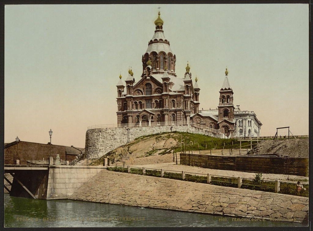 The image depicts a historic cathedral situated on elevated ground overlooking water. It features a large central dome with multiple smaller domes and crosses adorning the spires, indicative of Eastern Orthodox architecture commonly found in Russia or neighboring countries. The building is made up of intricate brickwork, stone facades, and ornate detailing around windows and entrances.

In front of the cathedral stands a fortified wall topped by a short fence, suggesting its significance as an important landmark possibly with historical defensive purposes. A bridge can be seen crossing over water in the foreground, connecting different parts of this area or providing access to it.

The overall color tone is muted with shades of grey and beige dominating the scene, implying that the photo may have been taken during a time when such colors were more prominent due to photographic techniques used at the time. The image carries historical weight as it reflects architectural styles from late 19th-century Russia or Finland (then known by its Russian name Helsinki).