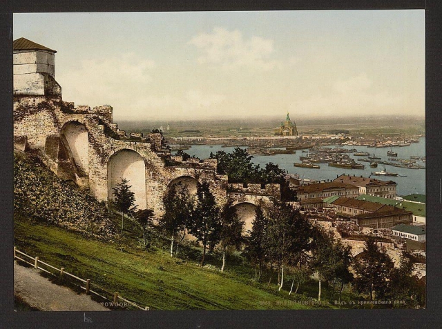 This image captures a panoramic view from what appears to be one of the Kremlin's ramparts, with Nizhny Novgorod in Russia visible below. The vantage point offers an expansive look at the cityscape and waterfront. Prominent features include aged stone arches part of the fortress walls that curve gracefully across the foreground. Beyond these structures lies a lush green landscape dotted with trees. A fence borders what seems to be a walking path beside this verdant area.

In the midground, several buildings in varying architectural styles line up along the waterfront. Their roofs and facades reflect different eras of construction, suggesting diversity over time. The water body is teeming with vessels of various sizes, indicating significant maritime activity or perhaps commerce. In the far background stands a distinctive dome structure atop an edifice near the horizon, possibly a church or another important building.

Above all this lies the sky, painted in soft hues that suggest either early morning light or late afternoon ambiance. The overall impression is one of historical richness and natural beauty intermingling with urban life.