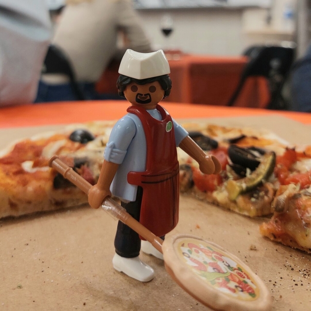 Pizzaiolo playmobil com avental vermelho, camisa azul, boina branca e com pá na mã. Atrás metade de uma pizza e ao fundo pessoas no restaurante