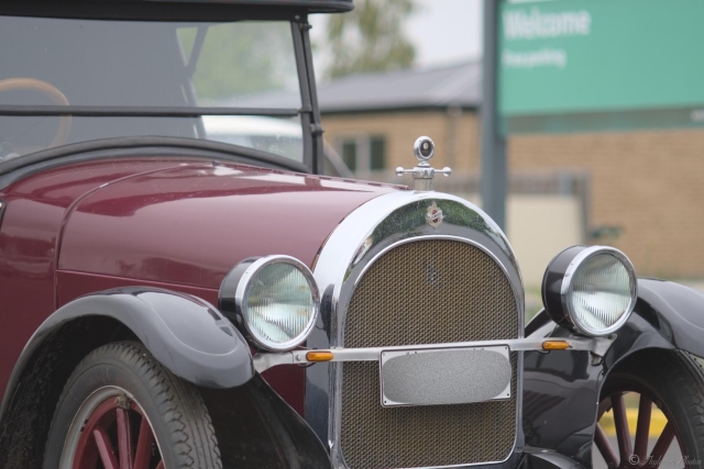 View of the front right hand side of an Oldmobile 2 seater vintage car with a soft roof, plenty of chrome, spoked wheels.  It has dark maroon paintwork with black wheel guards and trim.