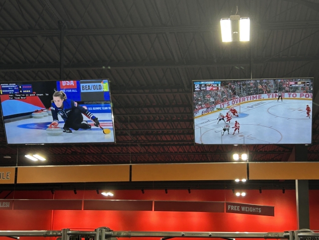 Two large screens display sports events in a gym. The left screen shows a female curler in action, while the right screen captures an NHL overtime hockey game between Minnesota and Detroit, currently tied at 3-3. The background features gym equipment