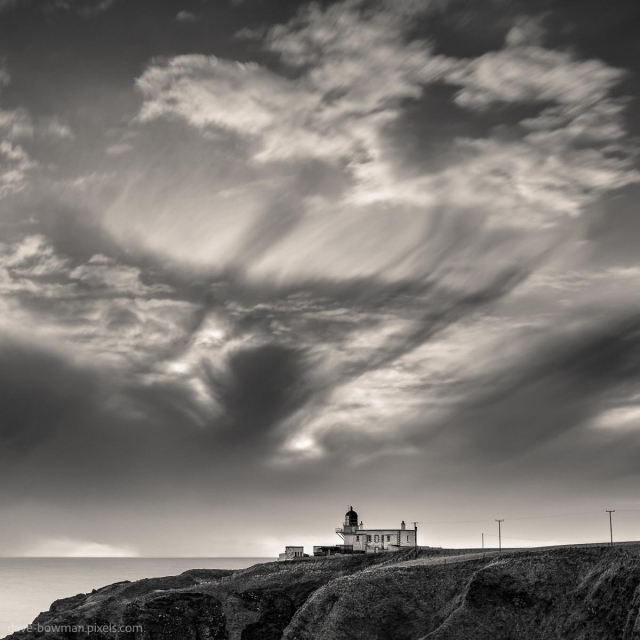 A photograph of Tod Head Lighthouse in Aberdeenshire, Scotland, set beneath dramatic clouds illuminated by early morning sunlight. The interplay of light and shadow creates strong contrasts, drawing attention to the rugged coastal landscape. The monochrome tones highlight the texture and depth of the clouds, giving a timeless quality to the scene and framing the iconic lighthouse against the dynamic sky.