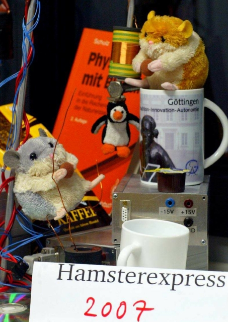 Titelseite „Hamsterexpress 2007“, Erstie-Zeitschrift FSR Physik Uni Göttingen. Zu sehen sind u.a. zwei kleine Plüschhamster, ein magnetischer Pinguin, eine Tasse „Göttingen Tradition – Innovation – Autonomie“, ein Netzteil, ein Physikbuch, eine Kaffeepackung, Geraffel.