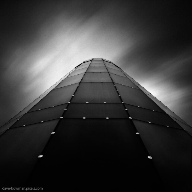 A photograph of a circular building in Amsterdam, Netherlands, highlighting its smooth, curving lines from an upward perspective. The semi-abstract composition draws attention to the building's flowing form, while streaked clouds drift across the sky, their softened appearance created by a long exposure, enhancing the moody atmosphere.