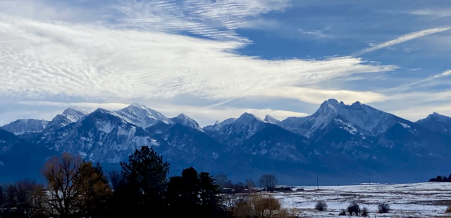 A wide shot that captures a mountain range’s impressive vista as jagged, snow and forest laden peaks rise dramatically out of the flat valley that’s still brown and white from winter. In the foreground to the left are trees. There are mixed, turbulent clouds over the mountains that the sun is illuminating.