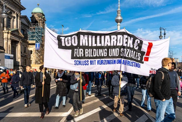 Das Foto von © Björn Obmann zeigt das Transparent mit dem Text: 100 Milliarden  € für Bildung und Soziales - Schluss mit Kürzungen und Aufrüstung!
Dahinter ist der Berliner Fernsehturm zu sehen sowie eine Reihe Demonstrant:*innen, die hinter dem Hochtransparent laufen.