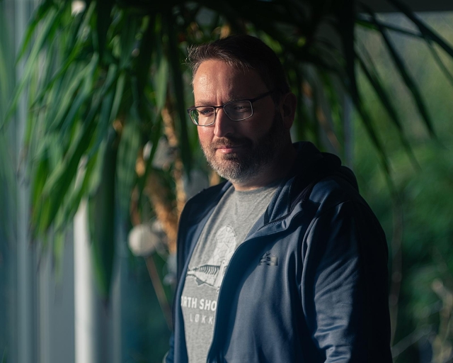 Male standing in a room in front of a palm tree and looking down to the ground