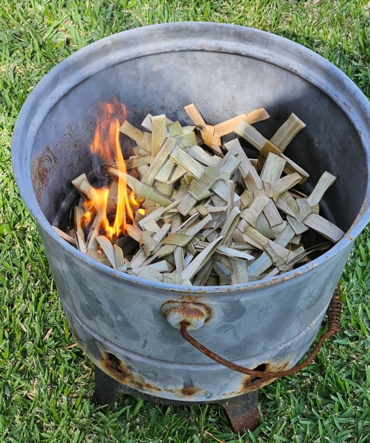 Pile of dried out palm crosses ignited in a small fire bowl sitting on a lawn