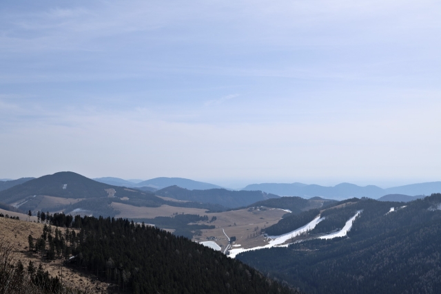 Blick über eine hügelige Landschaft. Im Vordergrund sieht man einen mit Nadelbäumen bewachsenen Hügel, dahinter liegt noch vereinzelt Schnee. In der Mitte unten kann man einen vereisten See erkennen. Der Himmel selbst ist blau mit Schleierwolken.