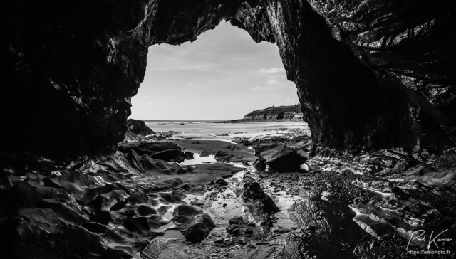 Photo monochrome en noir et blanc, prise depuis l'intérieur d'une grotte au pied d'une falaise. Par l'ouverture de la grotte, située à peu près au centre de la photo, on peut voir une plage de sable, découverte à marée basse avec à gauche sur la ligne d'horizon, la mer puis une falaise sur la droite. Le ciel est peu nuageux. Les côtés et le « toit » de la grotte sont dans la pénombre alors que le sol recouvert de rochers, polis par le mouvement de la marée, et de sable, bénéficie plus de la lumière extérieure.