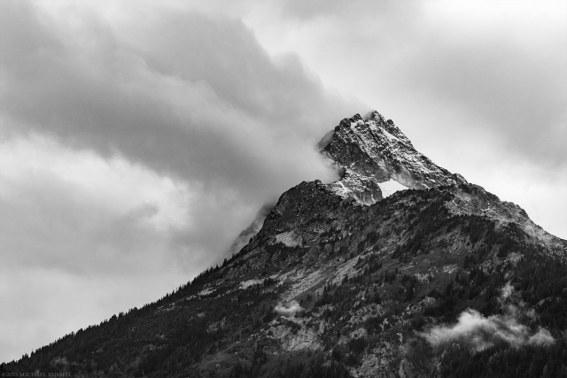 Black and white photo of a mountain peak with clouds appearing to stream off of the peak to the left with a cloud filled sky in the background.