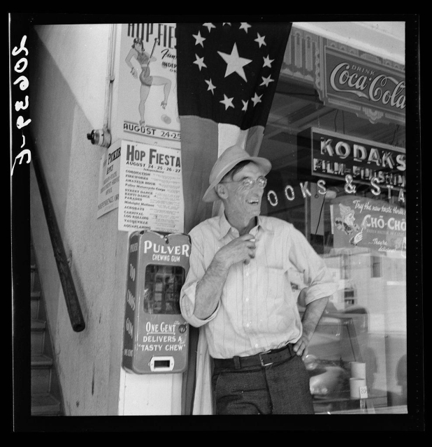 The image depicts a man standing next to various signs and posters in what appears to be an old-fashioned soda shop or diner. He is wearing glasses, a striped shirt with rolled-up sleeves, trousers, and he has his hand on the hip while smiling slightly as if posing for the photo. The background shows several items including: A Coca-Cola sign featuring its iconic logo with stars above it; A poster advertising "Hop Fiesta," an event scheduled for August 24-26 at a location called Covington Hall in Corvallis, Oregon; Another poster promoting a performance by the "Pulver Street Band" on October 15th, which is part of what seems to be a series with another date mentioned as well. There are also signs indicating other products available such as "Kodak Books & Stationery," and an advertisement for "Cho-Cho" chewing gum alongside a machine labeled 'Pulver' dispensing one cent chewing gum.

The overall atmosphere suggests this photo was taken during the late 1930s or early-to-mid-century, reflecting on daily life and community events of that era. The details in the image provide insights into consumer culture from past times as well as social activities within a small town setting.