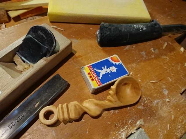 Handmade wood spoon. Knife, pack of matches, chisel and such on dirty work table.