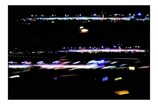 Fotografía abstracta desde la ventanilla del avión en la pista de despegue/aterrizaje. Sobre un fondo negro (es de noche) diferentes exposiciones de una de las terminales del aeropuerto desde lejos: vemos líneas de luces, jugando con varias exposiciones, el foco y algunas largas exposiciones algunas luces están desenfocadas, otras dibujan líneas al estar el avión ya moviéndose