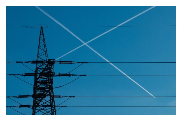 Foto im Querformat. Blick von unten auf die spitze eines Hochspannungsmastes. Diese ist links im Bild, fünf Leitungen verlaufen parallel nach rechts. Am blauen Himmel dahinter haben gerade zwei Flugzeuge ihren Weg gekreuzt und die Kondensstreifen bilden so ein weißes X bzw. ein Kreuz. In Kombination mit den Leitungen sieht es auch aus wie die schottische Flagge.