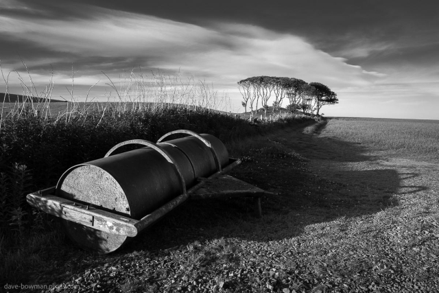 A photograph of a field roller on farmland in Crawton, Aberdeenshire, Scotland, shows the heavy, weathered machinery resting in the foreground. In the distance, a small copse of trees stands against the horizon. The long exposure creates soft, streaked clouds in the sky, adding a sense of movement to the otherwise still and open landscape. The monochrome tones emphasise the contrast between the textures of the land, machinery, and sky.