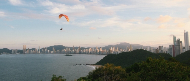 The image shows a paraglider soaring above a coastal landscape. Below, there's a large body of water, likely an ocean or sea, alongside a city with numerous skyscrapers. Hills or mountains frame the backdrop, and there is a lush, green foreground.