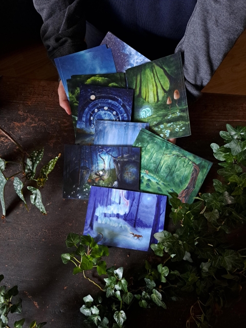 Hands holding a pile of postcards that are sliding to a table top surrounded by plants. The postcards have different colorful illustrations inspired by nordic mythology such as a fox creating northern lights and a moose carrying the sun between his antlers.