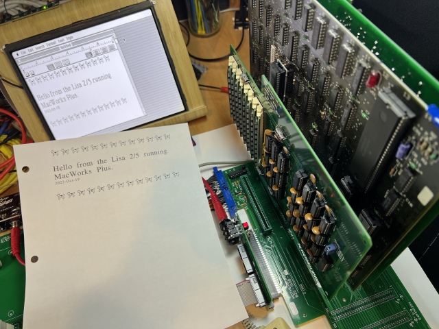 A messy desk with a pile of large green circuit boards to the right of a #AppleLisaClone. In the back is a makeshift display showing a MacWrite document. In the foreground is a letter sized printed page from MacWrite that says “Hello from the Lisa 2/5 running MacWorks Plus” along with many cowdogs at the top and bottom of the page (moof!).