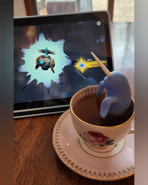 A pink teacup, with a smiling blue narwhal tea infuser relaxing in the hot tea, rests in front of a tablet that displays the "Cashflow Supervision" weapon from Genshin Impact's wishing gacha. This is the signature weapon of a character who likes drinking tea.