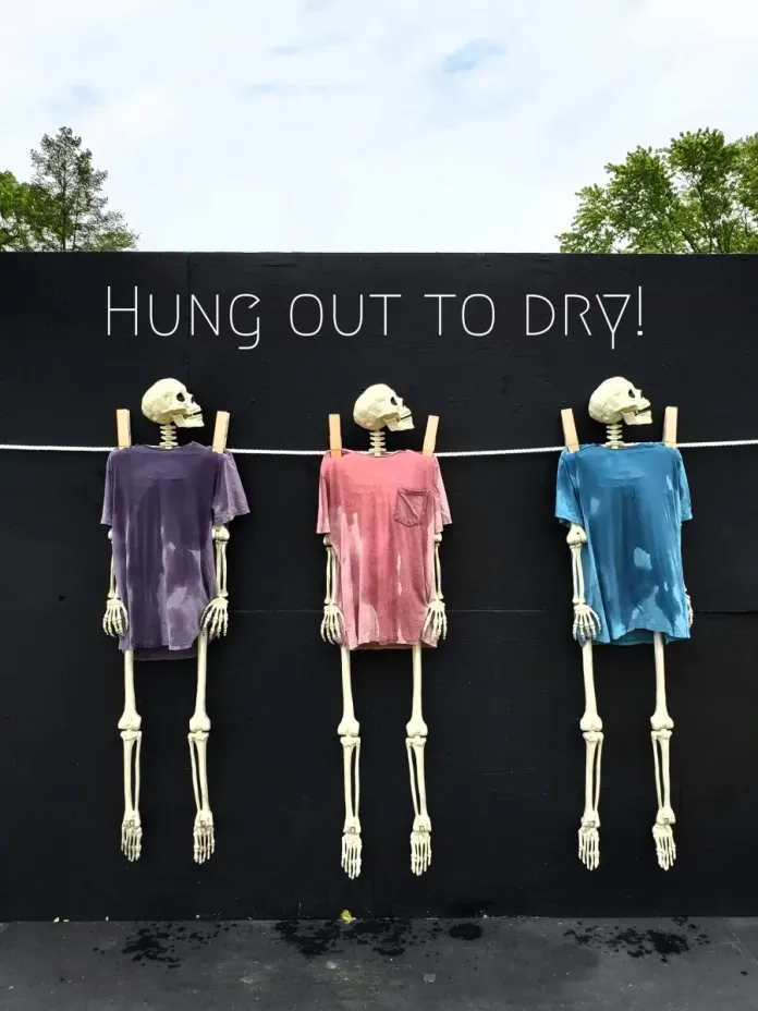 "Hung Out to Dry" by ItSkeletal:Three skeletons hang from a clothesline, each wearing a soaked T-shirt in different colors—purple, red, and blue—pinned up like laundry. The phrase "Hung Out to Dry!" is painted above them. A witty visual metaphor about exposure, abandonment, or simply a playful dark humor take on everyday routines.