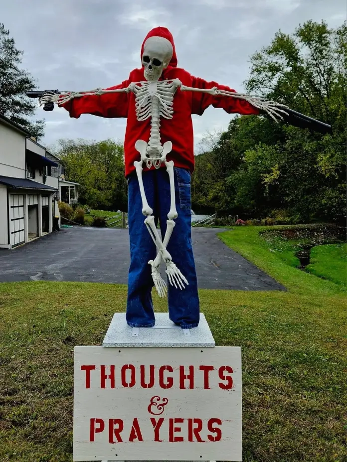 "Thoughts & Prayers" by ItSkeletal – A skeleton dressed in a red hoodie, posed like a scarecrow, holds guns in both hands while standing on a sign that reads "Thoughts & Prayers." A bold and unsettling commentary on the emptiness of words in response to gun violence.